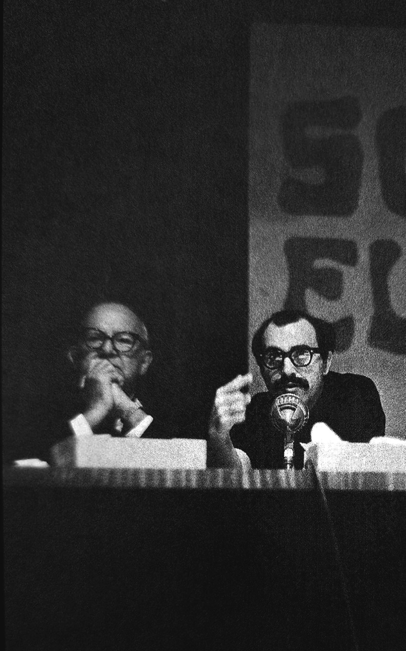 Villanueva en una conferencia, rostro iluminado frente al micrófono